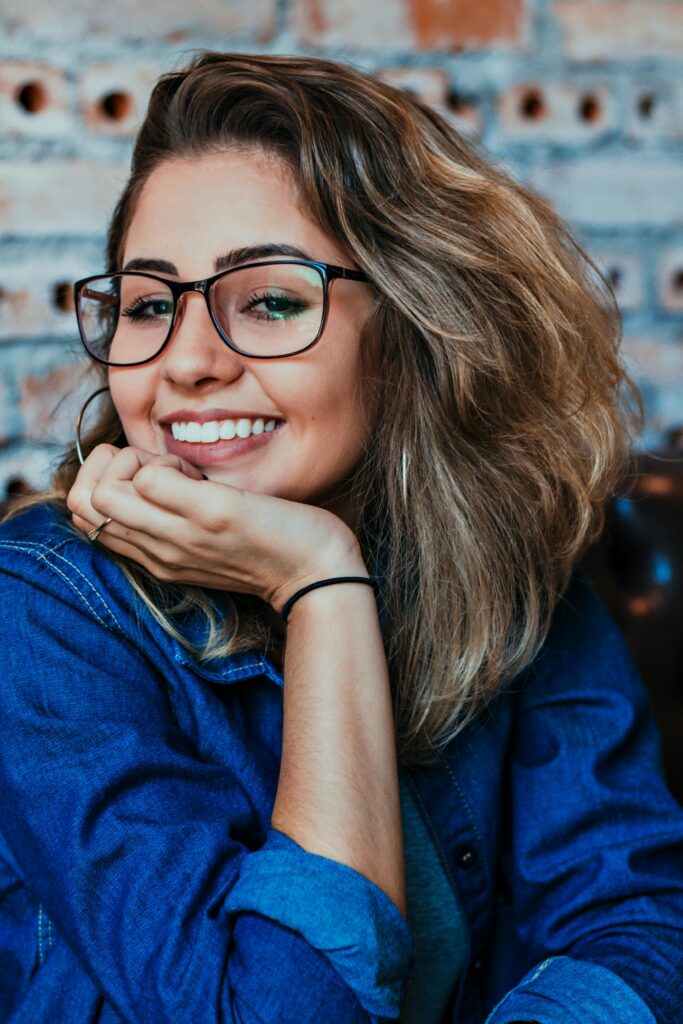 pexels-photo-1130626-1130626 A young woman wearing glasses and denim, smiling in an indoor setting.