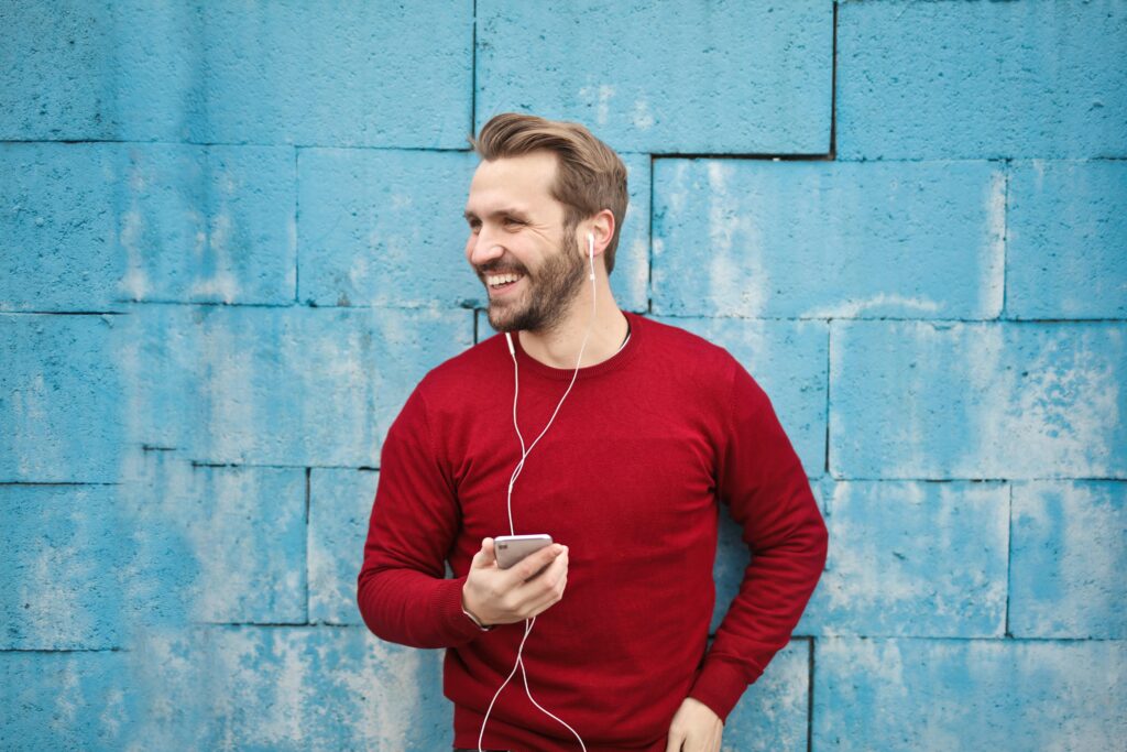 pexels-photo-846741-846741 A joyful man in a red sweater smiles while listening to music on his smartphone against a blue brick wall.